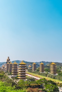 A series of traditional pagodas with golden rooftops are set against a backdrop of rolling green hills. A large Buddha statue is visible at the end of a pathway lined with greenery and landscaping.