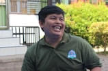A person with short dark hair is laughing joyfully while wearing a green collared shirt with a badge. The background includes leafy greenery and a staircase with a railing, suggesting an outdoor setting.
