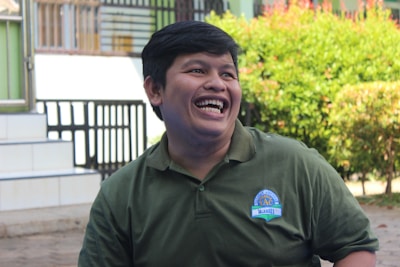 A person with short dark hair is laughing joyfully while wearing a green collared shirt with a badge. The background includes leafy greenery and a staircase with a railing, suggesting an outdoor setting.
