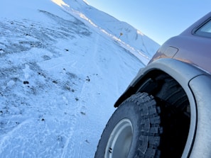 A close-up of a car tire on a rocky trail with snow-capped peaks in the background.