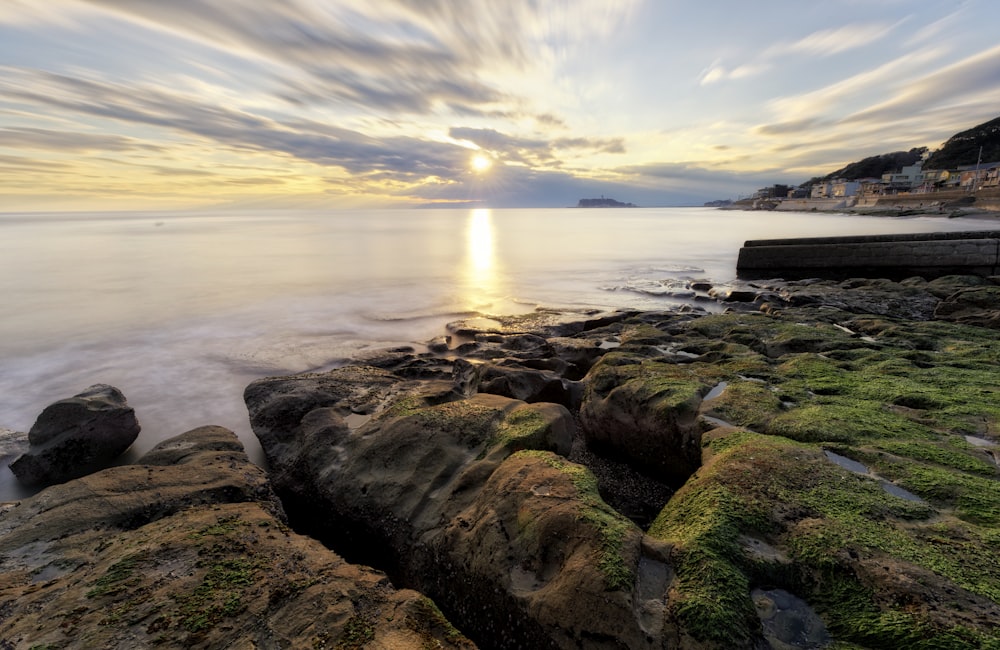 the sun is setting over the ocean with rocks in the foreground