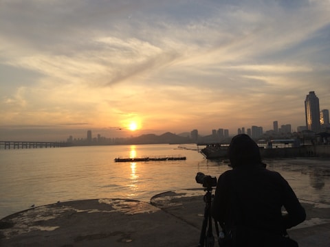 A person is photographing a serene waterfront during a golden sunset. Silhouetted against the sky, the individual uses a camera on a tripod. Distant skyscrapers and a long bridge span across the horizon, reflecting softly on the water's surface. The sky is painted with warm hues of orange and yellow.