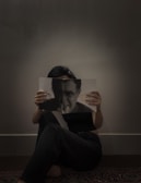 A person sits on the floor against a plain wall, holding a magazine or book that partially covers their face. The image on the cover features a monochrome portrait that aligns with the person's eyes, creating a visual illusion.