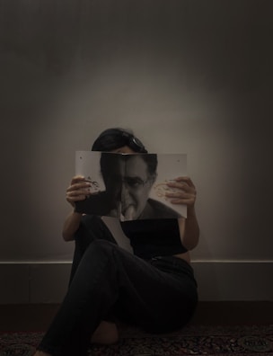 A person sits on the floor against a plain wall, holding a magazine or book that partially covers their face. The image on the cover features a monochrome portrait that aligns with the person's eyes, creating a visual illusion.