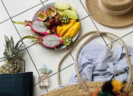 On a white tiled floor, a vibrant platter of tropical fruits including dragon fruit, passion fruit, strawberries, kiwi, and mango is artfully arranged. Nearby, a pineapple and a pastel-colored perfume bottle are placed next to a straw sun hat and a woven beach bag containing a blue and white striped fabric.