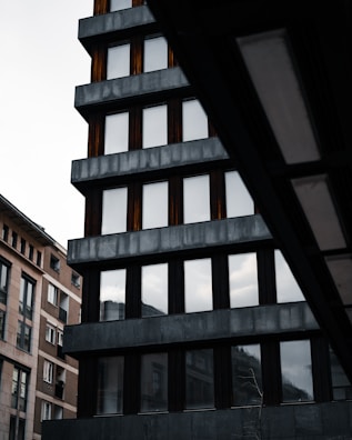 A modern building with a grid of dark, reflective windows and a concrete exterior. The architectural design features clean, straight lines and a strong vertical emphasis. Adjacent buildings are visible, showing contrasting materials and older designs.