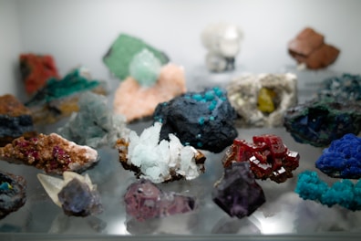A diverse collection of colorful minerals and crystals displayed on a reflective surface, featuring a wide array of shapes and textures, with hues ranging from deep reds and blues to greens, browns, and whites.
