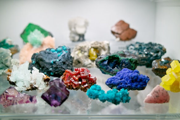 A variety of colorful mineral samples displayed on a reflective surface. The minerals are of different shapes, sizes, and colors, including striking reds, blues, greens, yellows, and purples.