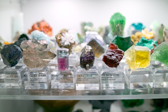 A collection of various colorful mineral crystals displayed on clear stands, each labeled with their names and origins. The crystals vary in color, shape, and texture, including vibrant hues of pink, green, yellow, red, and purple. The background shows a range of other neatly arranged specimens.