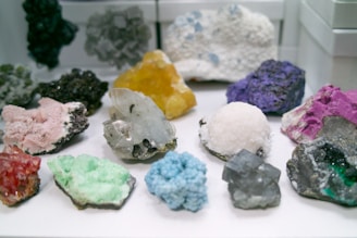 A collection of various colorful mineral specimens displayed on a white surface. The minerals vary in shape and texture, showcasing hues such as pink, green, blue, purple, red, yellow, and white.