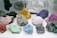 A collection of various colorful mineral specimens displayed on a white surface. The minerals vary in shape and texture, showcasing hues such as pink, green, blue, purple, red, yellow, and white.