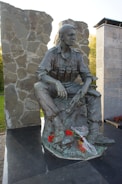 A bronze statue of a soldier seated with a rifle laid across his lap, amidst a memorial setting with stone walls and inscriptions nearby. Red flowers and a piece of fabric are placed at the base of the statue.