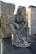 A bronze statue of a soldier seated with a rifle laid across his lap, amidst a memorial setting with stone walls and inscriptions nearby. Red flowers and a piece of fabric are placed at the base of the statue.