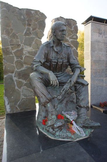 A bronze statue of a soldier seated with a rifle laid across his lap, amidst a memorial setting with stone walls and inscriptions nearby. Red flowers and a piece of fabric are placed at the base of the statue.