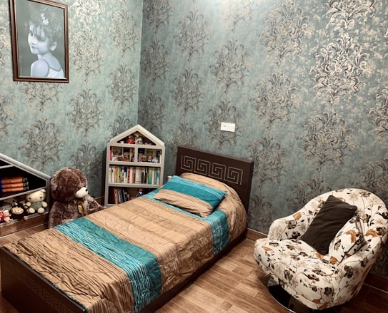 A cozy and well-decorated bedroom features a single bed with a brown and teal bedspread, a bookshelf shaped like a house containing various books and toys, and a plush teddy bear seated beside the bed. The dark brown flooring complements the ornate wallpaper with floral patterns. A comfortable armchair with floral upholstery sits beside the bed, adding to the room's inviting atmosphere. A large framed portrait of a child hangs on one of the walls.