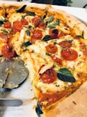 A delicious pizza topped with melted cheese, juicy cherry tomatoes, and fresh basil leaves. A slice has been removed, revealing the golden, crispy crust. A pizza cutter is partially visible on a metal tray.