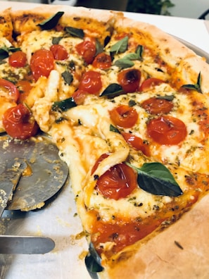 A delicious pizza topped with melted cheese, juicy cherry tomatoes, and fresh basil leaves. A slice has been removed, revealing the golden, crispy crust. A pizza cutter is partially visible on a metal tray.