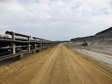 A vast, industrial landscape featuring a long conveyor belt extending into the distance on the left side, while the right side shows a large excavated dirt pathway. A construction vehicle is visible in the background. The sky is overcast, creating a somber and utilitarian mood.