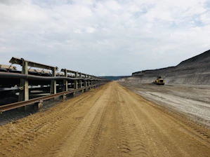 A vast, industrial landscape featuring a long conveyor belt extending into the distance on the left side, while the right side shows a large excavated dirt pathway. A construction vehicle is visible in the background. The sky is overcast, creating a somber and utilitarian mood.