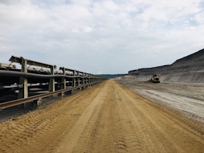 A vast, industrial landscape featuring a long conveyor belt extending into the distance on the left side, while the right side shows a large excavated dirt pathway. A construction vehicle is visible in the background. The sky is overcast, creating a somber and utilitarian mood.