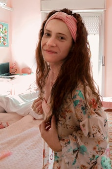 A woman with long hair wearing a floral patterned robe and a pink headband is in a brightly lit bedroom. The room has a bed with white linens, a desk with a laptop, and a pink chair. A colorful artwork is visible on the wall.