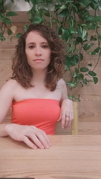 A person with curly hair is seated at a wooden table wearing a strapless coral dress. The background is adorned with hanging green plants and wooden paneling.