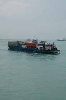 A barge is carrying several vehicles across a calm body of water. The sky is overcast, and there are a few seagulls flying around. In the distance, there is another vessel visible on the horizon.