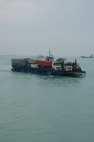 A barge is carrying several vehicles across a calm body of water. The sky is overcast, and there are a few seagulls flying around. In the distance, there is another vessel visible on the horizon.
