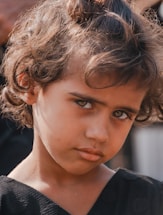 A young child with curly hair gazes directly at the camera, creating a thoughtful and somewhat intense expression. The child is wearing a dark top and is positioned against a blurred background that subtly suggests an outdoor setting.