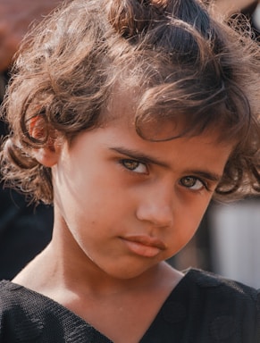 A young child with curly hair gazes directly at the camera, creating a thoughtful and somewhat intense expression. The child is wearing a dark top and is positioned against a blurred background that subtly suggests an outdoor setting.