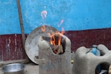 A traditional clay stove is in use, with firewood burning brightly inside. The background consists of a textured blue wall with a red strip at the base. Nearby, there are various household items, including a metal bowl on the ground and a plastic bottle within a clay holder.