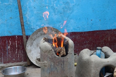 A traditional clay stove is in use, with firewood burning brightly inside. The background consists of a textured blue wall with a red strip at the base. Nearby, there are various household items, including a metal bowl on the ground and a plastic bottle within a clay holder.