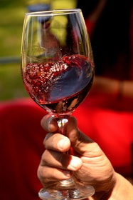 A hand is holding a wine glass filled with red wine, creating a circular motion that causes the wine to swirl and create bubbles. The background is softly blurred with hints of green and red, suggesting an outdoor setting.