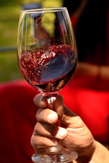 A hand is holding a wine glass filled with red wine, creating a circular motion that causes the wine to swirl and create bubbles. The background is softly blurred with hints of green and red, suggesting an outdoor setting.