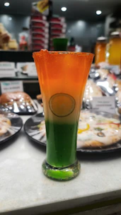 A tall, elegant glass filled with a layered beverage featuring vibrant green, white, and orange hues, resembling the Indian national flag. A lemon slice is submerged in the white section, adding a decorative touch. The background shows blurred images of various pastries and drinks.