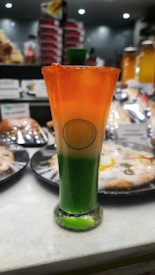 A tall, elegant glass filled with a layered beverage featuring vibrant green, white, and orange hues, resembling the Indian national flag. A lemon slice is submerged in the white section, adding a decorative touch. The background shows blurred images of various pastries and drinks.
