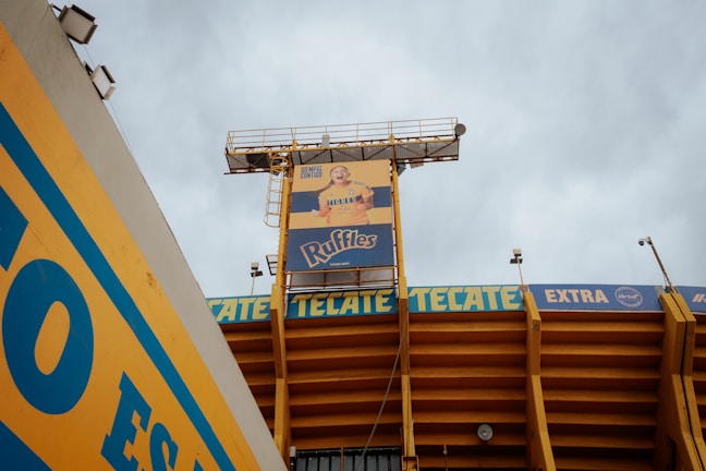 A stadium structure with a large advertisement for Ruffles featuring a person wearing a yellow shirt is prominently displayed. The stadium is painted in yellow and blue, with the brand names 'Tecate' and 'Extra' visible along the upper sections of the structure.