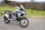 A modern adventure motorcycle is parked on a paved path with a grassy field and bare trees in the background. The motorcycle features knobby tires, protective guards, and a windscreen, indicating it is designed for off-road or touring use. The environment appears calm and quiet, with the scene set against a backdrop of rolling hills and scattered leafless trees.