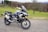 A modern adventure motorcycle is parked on a paved path with a grassy field and bare trees in the background. The motorcycle features knobby tires, protective guards, and a windscreen, indicating it is designed for off-road or touring use. The environment appears calm and quiet, with the scene set against a backdrop of rolling hills and scattered leafless trees.