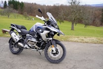 A modern adventure motorcycle is parked on a paved path with a grassy field and bare trees in the background. The motorcycle features knobby tires, protective guards, and a windscreen, indicating it is designed for off-road or touring use. The environment appears calm and quiet, with the scene set against a backdrop of rolling hills and scattered leafless trees.