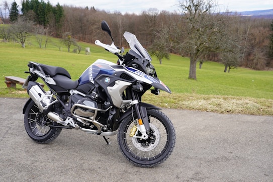 A modern adventure motorcycle is parked on a paved path with a grassy field and bare trees in the background. The motorcycle features knobby tires, protective guards, and a windscreen, indicating it is designed for off-road or touring use. The environment appears calm and quiet, with the scene set against a backdrop of rolling hills and scattered leafless trees.