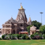 A large, intricately designed Hindu temple with multiple domes and ornate carvings stands majestically against a clear blue sky. The structure is made of beige stone with golden accents on its spires. Lush green gardens surround the temple.