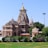 A large, intricately designed Hindu temple with multiple domes and ornate carvings stands majestically against a clear blue sky. The structure is made of beige stone with golden accents on its spires. Lush green gardens surround the temple.