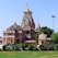 A large, intricately designed Hindu temple with multiple domes and ornate carvings stands majestically against a clear blue sky. The structure is made of beige stone with golden accents on its spires. Lush green gardens surround the temple.