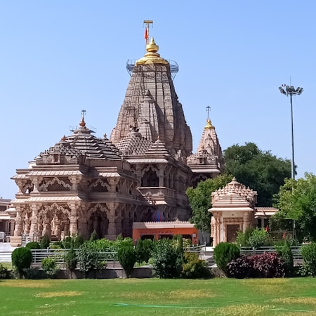 A large, intricately designed Hindu temple with multiple domes and ornate carvings stands majestically against a clear blue sky. The structure is made of beige stone with golden accents on its spires. Lush green gardens surround the temple.