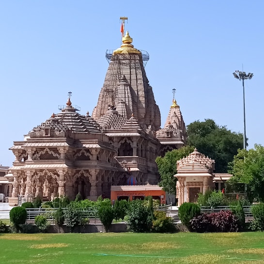 A large, intricately designed Hindu temple with multiple domes and ornate carvings stands majestically against a clear blue sky. The structure is made of beige stone with golden accents on its spires. Lush green gardens surround the temple.