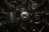 A close-up view of various black metal screws and fasteners, with one silver screw prominently in focus. The background is blurred, creating a dark, industrial atmosphere.