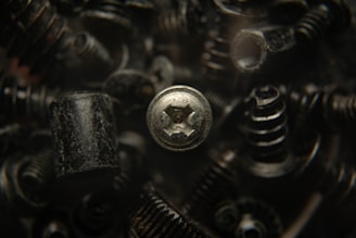 A close-up view of various black metal screws and fasteners, with one silver screw prominently in focus. The background is blurred, creating a dark, industrial atmosphere.