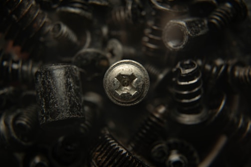 A close-up view of various black metal screws and fasteners, with one silver screw prominently in focus. The background is blurred, creating a dark, industrial atmosphere.