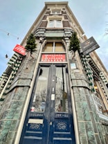 A tall, ornate building facade with an entrance featuring a dark, intricately designed wooden door. The architecture includes green-tiled walls and windows with white decorative elements. Several signs, including one for 'Mosselen', are visible along with small evergreen trees for decoration.